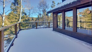 Una terraza de madera con suelo cubierto de nieve, grandes ventanales y vistas a árboles y montañas.