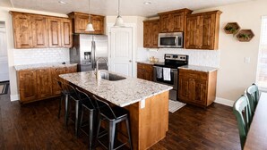 A kitchen with wooden cabinets, a stainless steel refrigerator, a microwave, an oven, and a granite island with bar stools. - Sleeps 16 • Family Retreat Near BYU and UVU• Spacious 6BR Home,perfect for group (Orem)