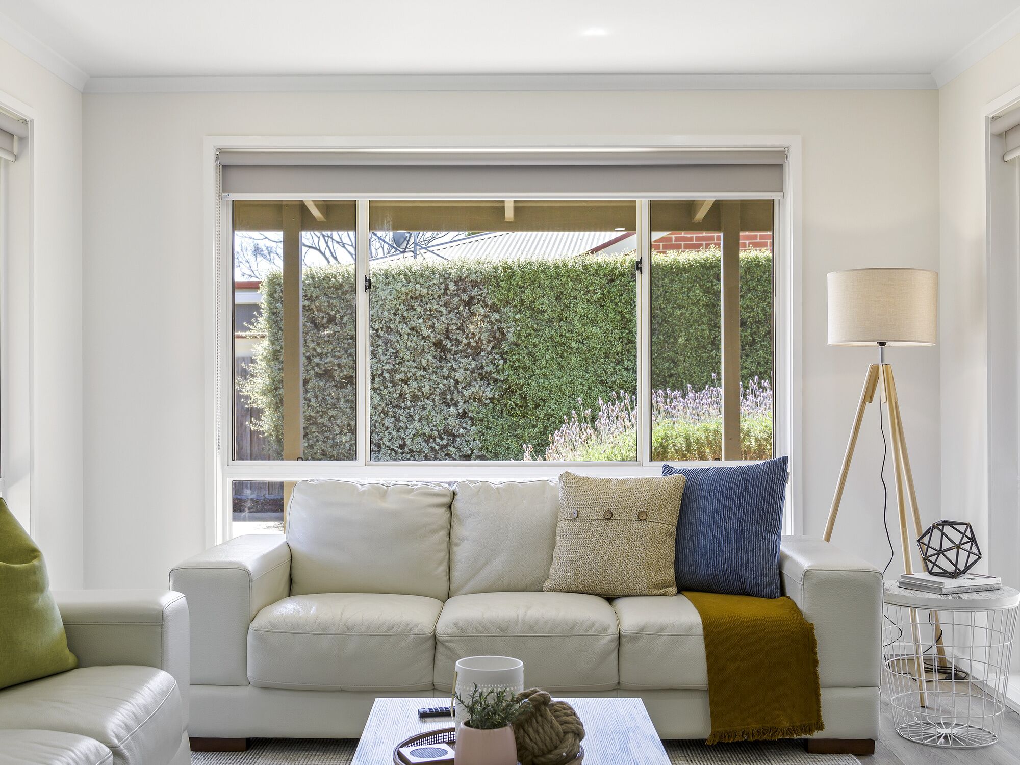 Una sala de estar moderna con un sofá de cuero blanco, una mesa de centro con objetos decorativos, una lámpara de pie y vistas a un seto a través de amplias ventanas.