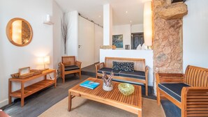 A modern living room with a stone accent wall, wooden furniture, and a view of the kitchen. - JOIVY Elegant Beach Studio (Portinho da Arrábida, Azeitão)