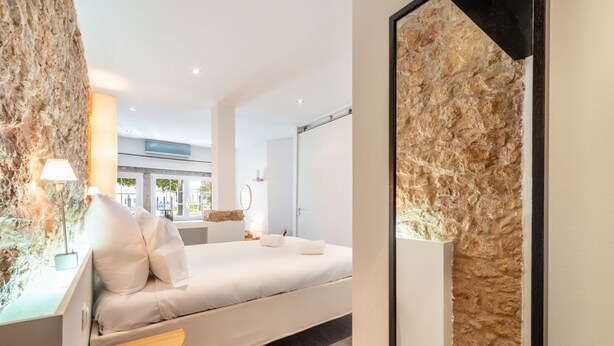 A modern hotel room with a large bed, a wooden side table, a stone accent wall, and a view of the outdoors. - JOIVY Elegant Beach Studio (Portinho da Arrábida, Azeitão)