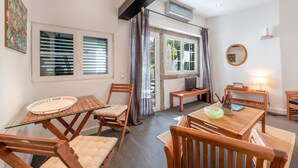 A cozy room with a glass table, wooden chairs, a TV, and a window with blinds. - JOIVY Elegant Beach Studio (Portinho da Arrábida, Azeitão)