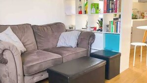 Ein Wohnzimmer mit grauem Sofa, einem schwarzen Hocker und einem weißen Tisch. Ein Bücherregal zeigt Bücher und Dekorationselemente.
