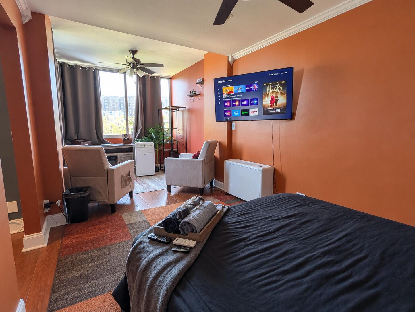 A hotel room with a bed, a TV mounted on the wall, a desk with a chair, and a view of the outside through a window.