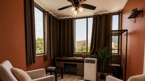 Ein Wohnzimmer mit zwei beigefarbenen Sesseln, einem kleinen Tisch, einem Deckenventilator, einem Fenster mit Vorhängen, einem Heizkörper und Blick auf ein Gebäude außerhalb.