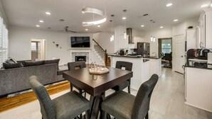 Uma sala de estar moderna e integrada, com uma mesa de jantar, sofás cinzas e uma cozinha ao fundo.