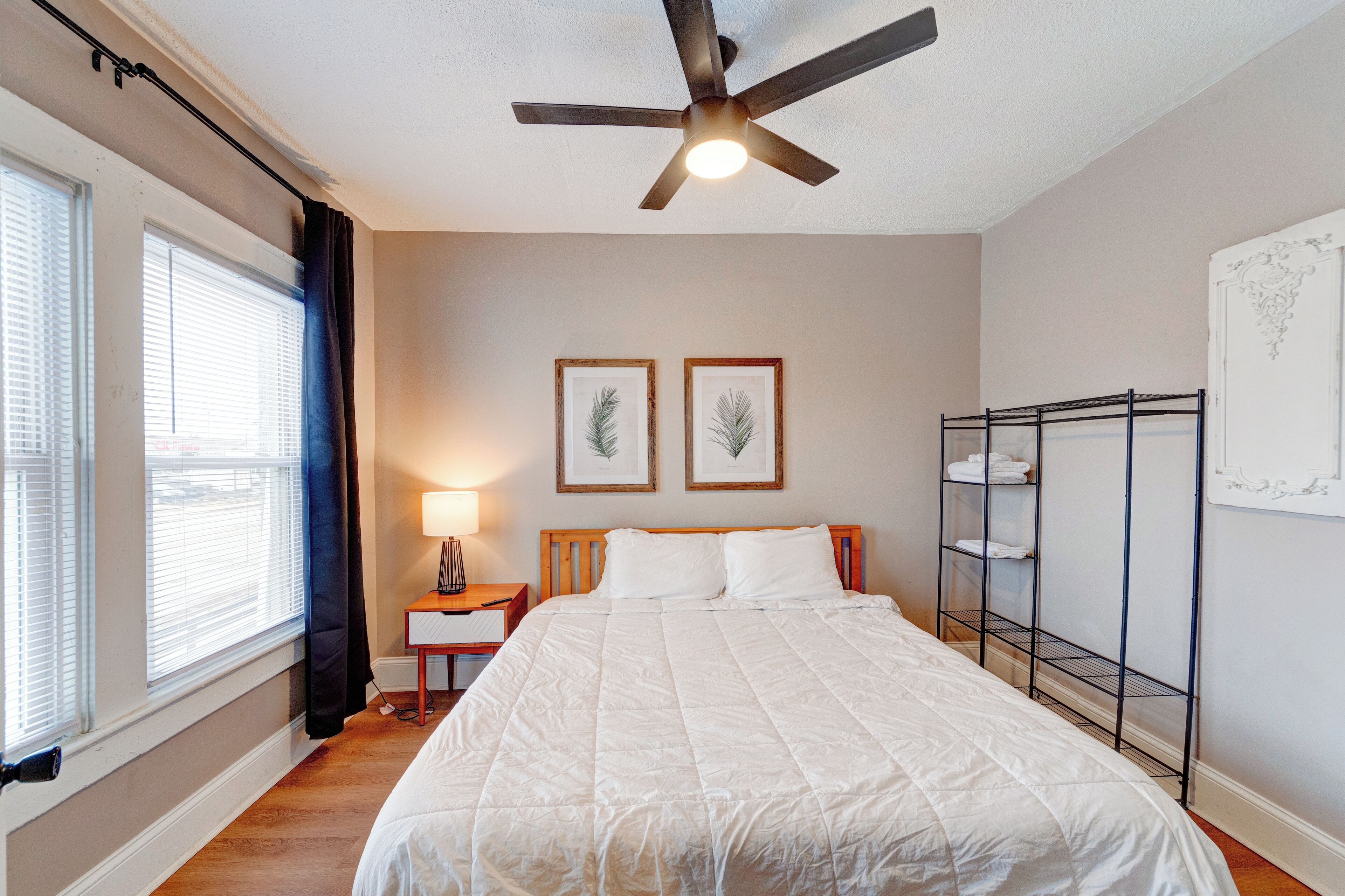 A bedroom with a bed, a ceiling fan, a nightstand, a lamp, framed pictures on the wall, and a metal shelf unit.