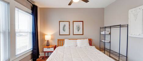 A bedroom with a bed, a ceiling fan, a nightstand, a lamp, framed pictures on the wall, and a metal shelf unit.