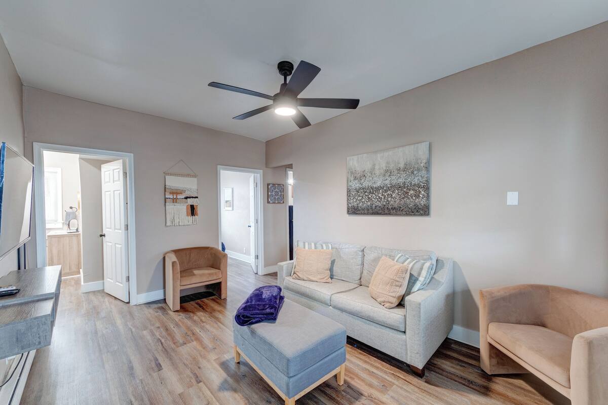 A spacious living room with a ceiling fan, a light-colored sofa, a wooden floor, and a door leading to another room.