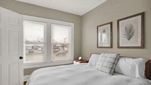A bedroom with a large bed, two windows, a nightstand, and two framed botanical prints on the wall. - The village (Indianapolis)