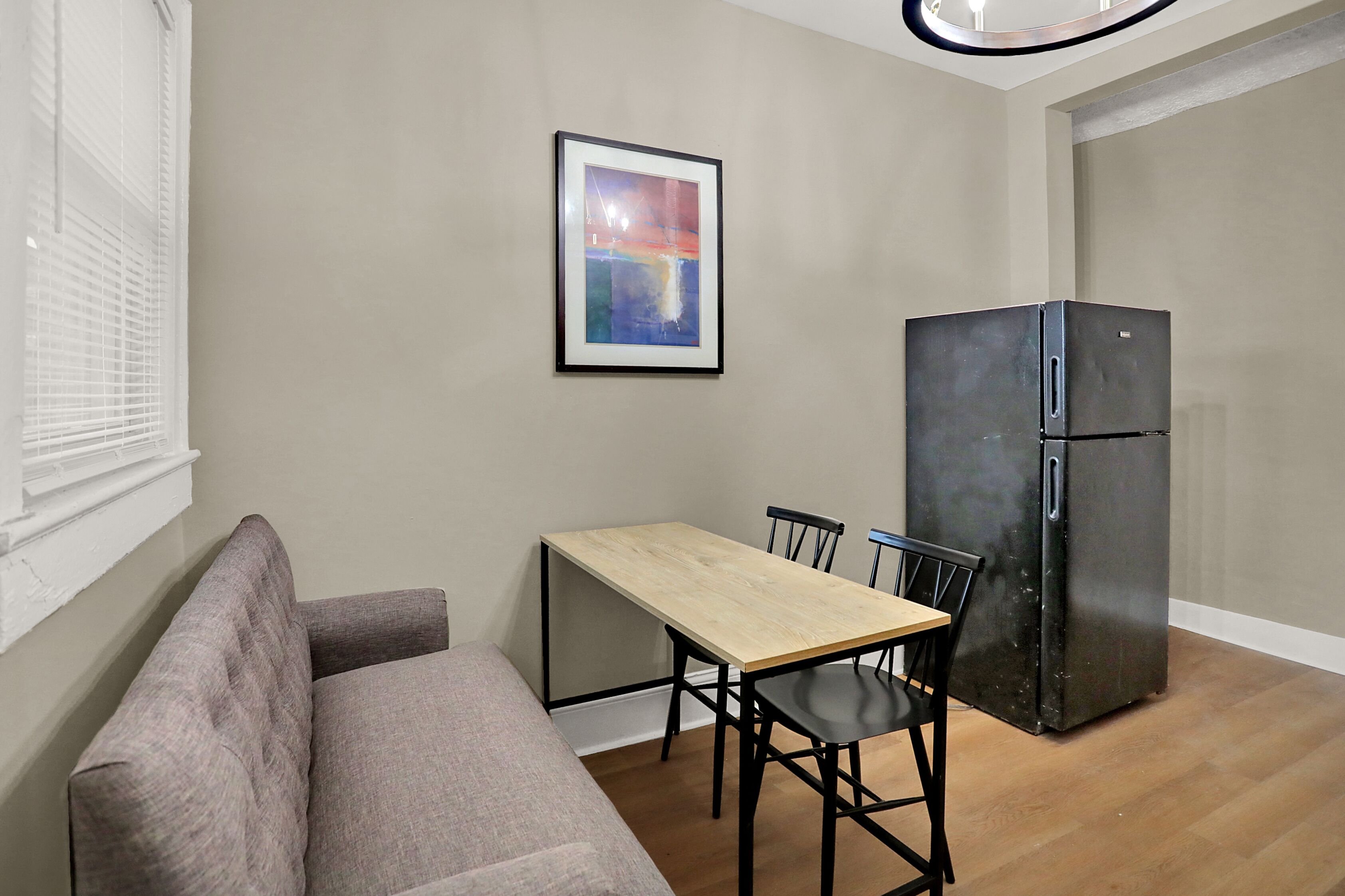 A compact living space with a sofa, a dining table with chairs, a black refrigerator, and a framed abstract painting on the wall.