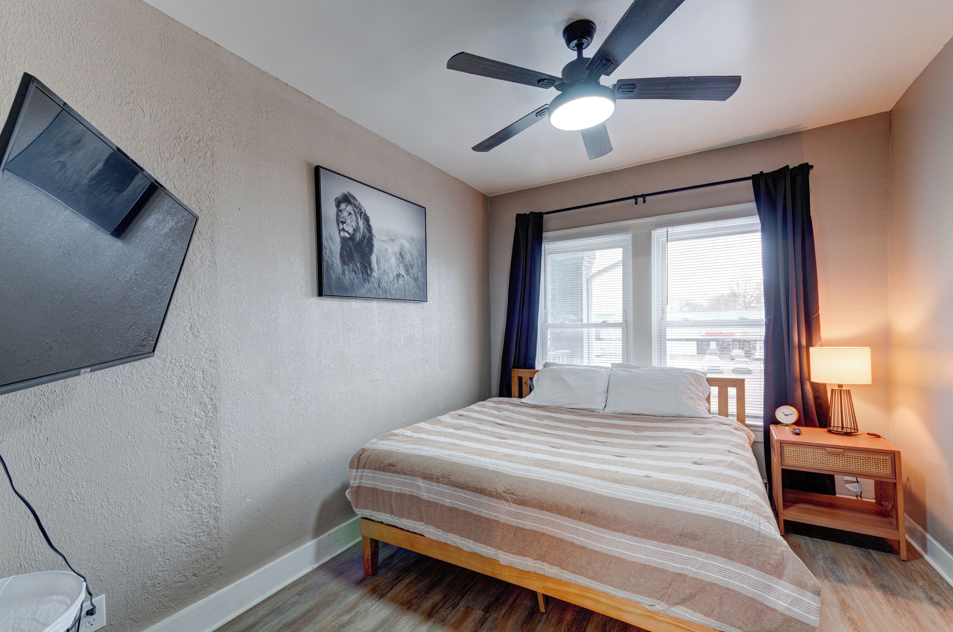 A hotel room with a bed, a ceiling fan, a wall-mounted TV, a window with curtains, and a bedside table with a lamp.