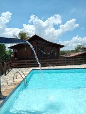 Une piscine avec une fontaine, une terrasse en bois et, au fond, un bâtiment doté d’un toit recouvert de tuiles.