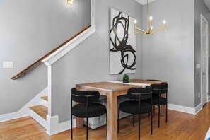 Un comedor moderno con una mesa de madera y sillas negras, una escalera con barandillas blancas y un cuadro abstracto contemporáneo en la pared.