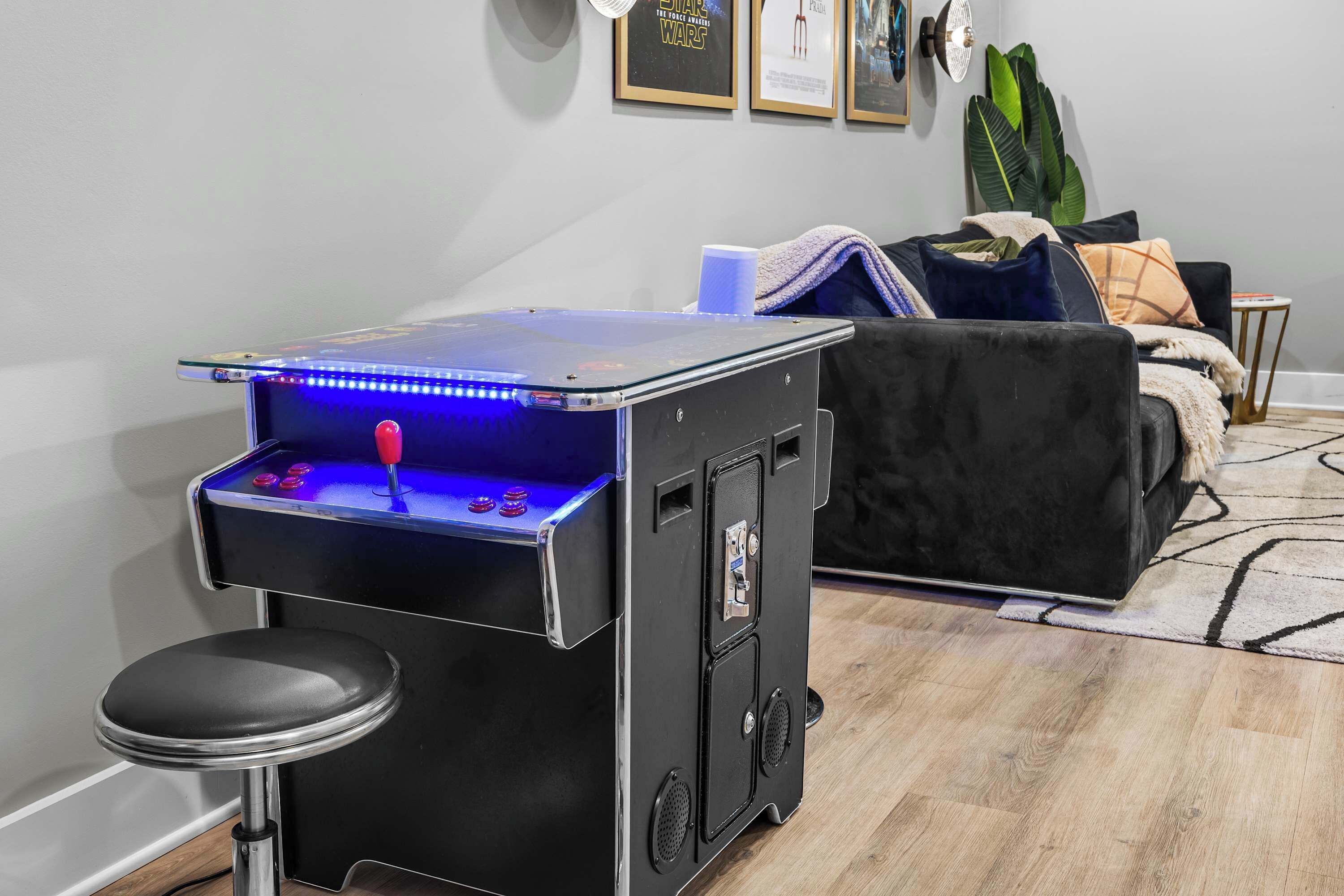 Quarto com uma máquina de jogos eletrônicos pequena, um sofá preto, uma mesa de centro e uma planta.