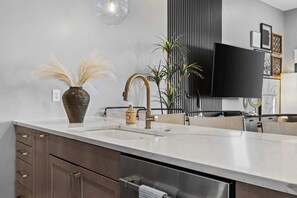 Una cocina moderna con encimera blanca, pileta de acero inoxidable, grifo dorado y un jarrón con plantas secas.