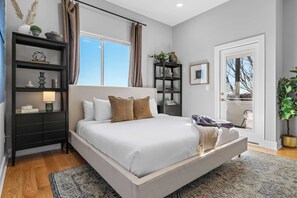 A modern bedroom with a large bed, a nightstand, a bookshelf, and a view of the outdoors through a glass door.