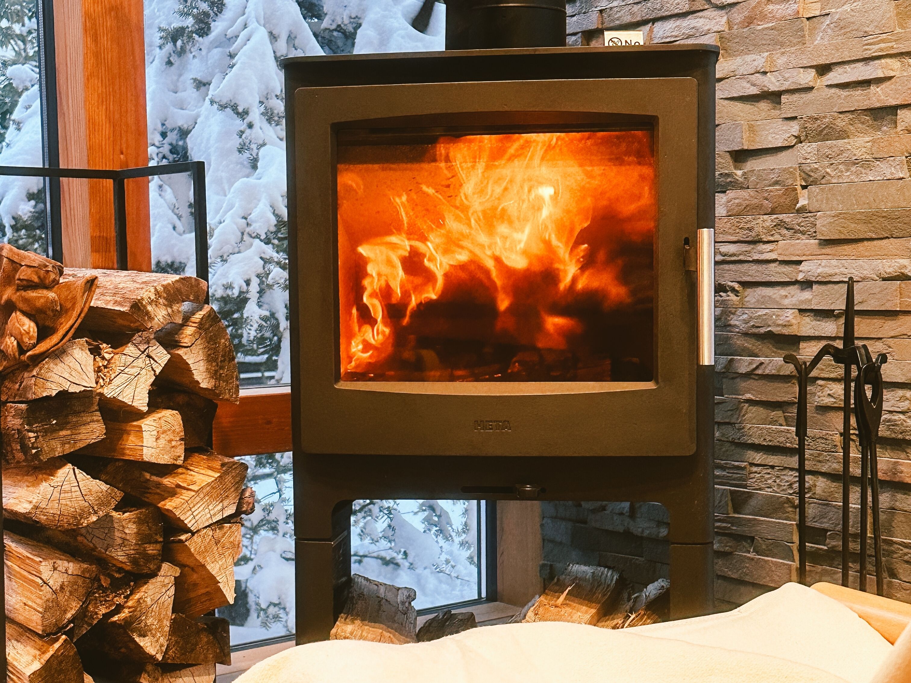 A wood-burning stove with a stone wall backdrop and a stack of firewood.
