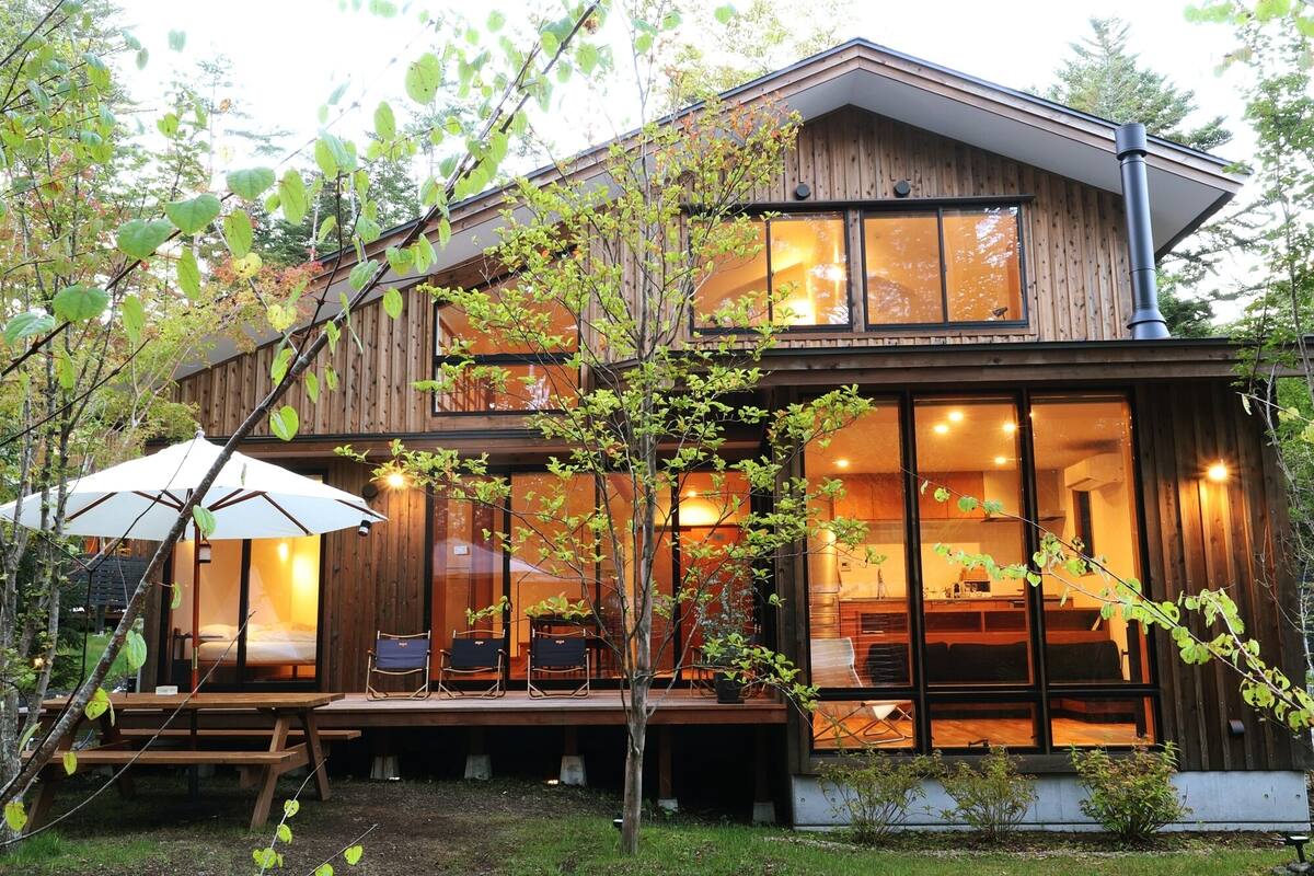 A two-story wooden house with large windows, a deck with chairs, and a patio umbrella.