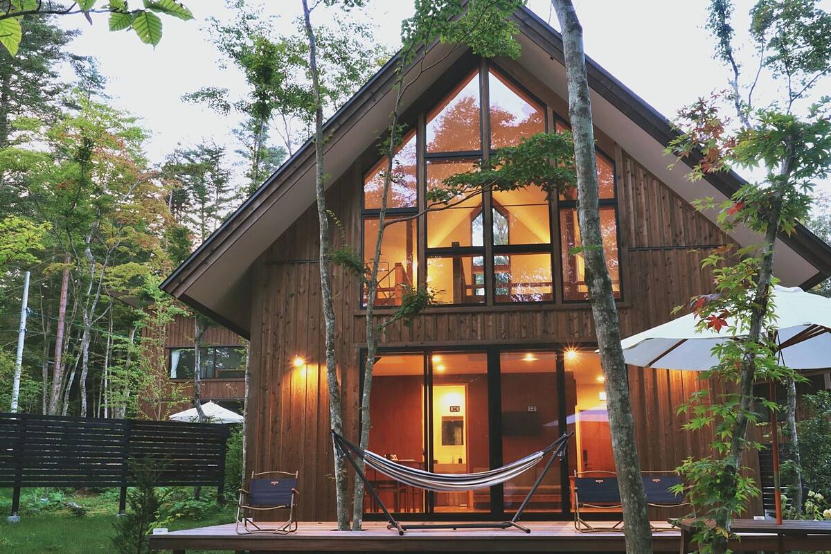 A two-story wooden cabin with large windows, a hammock, and outdoor seating.