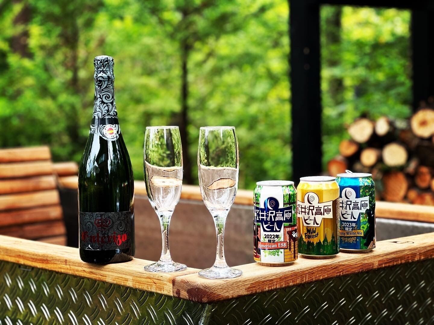 A bottle of wine, two glasses, and three cans of beer on a wooden table with a forest background.