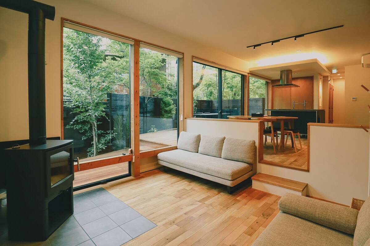 A modern living room with a sofa, a dining table, and a kitchen area visible through large windows.
