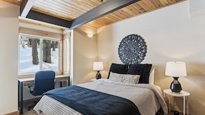 Une chambre à coucher comprenant un lit, deux lampes de chevet, un élément décoratif mural, un bureau avec une chaise et une fenêtre donnant sur des arbres et de la neige.