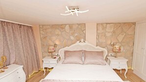 A bedroom with a stone wall, a white bed, two bedside tables with lamps, and a white wardrobe.