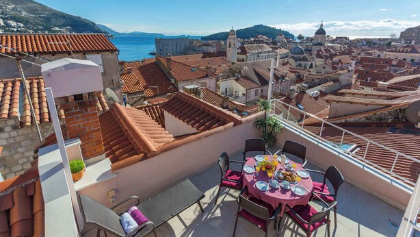 A rooftop terrace with a table set for dining, overlooking a coastal cityscape with terracotta rooftops and historic buildings.
