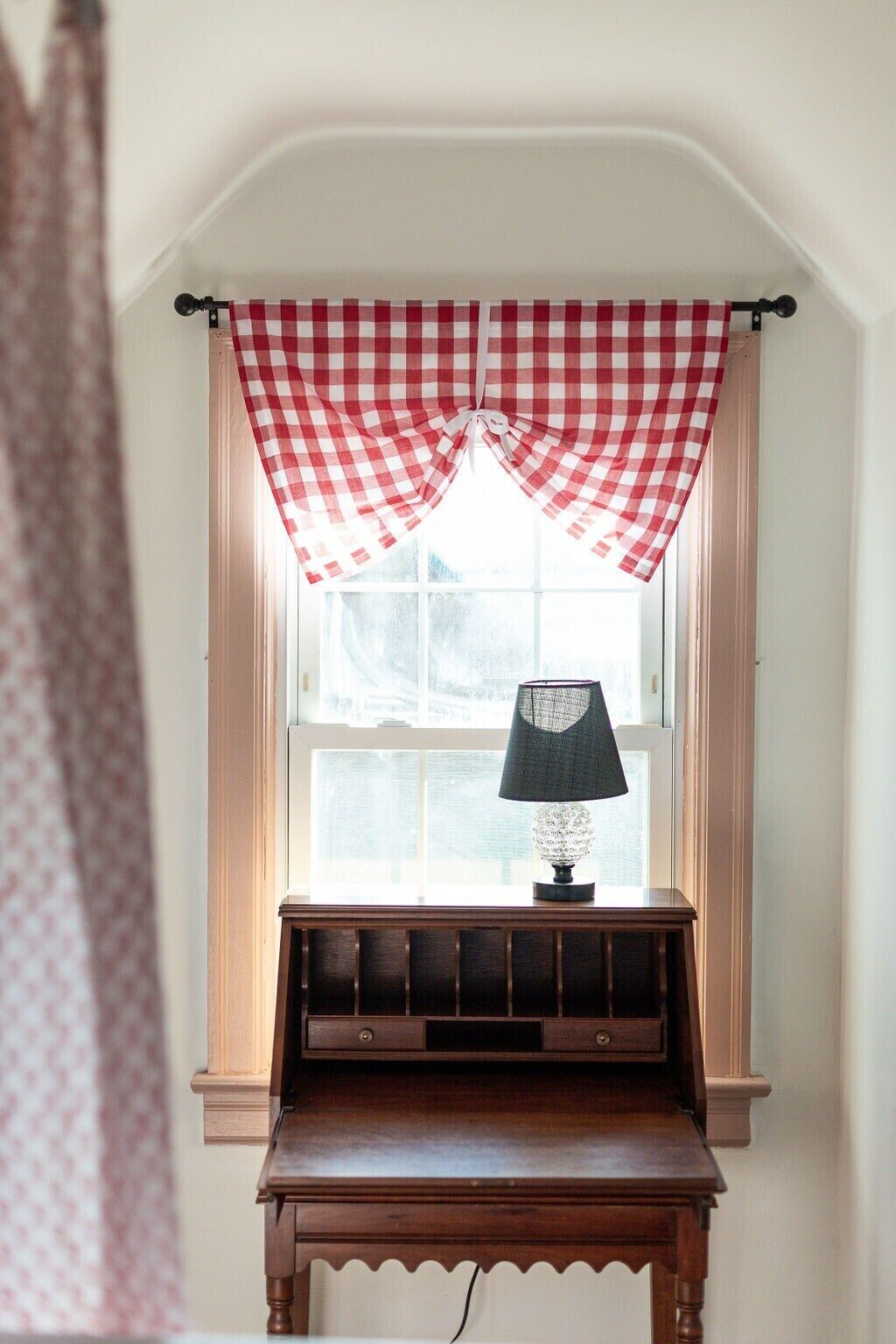 A room with a window, a lamp on a wooden table, and a red and white checkered curtain.
