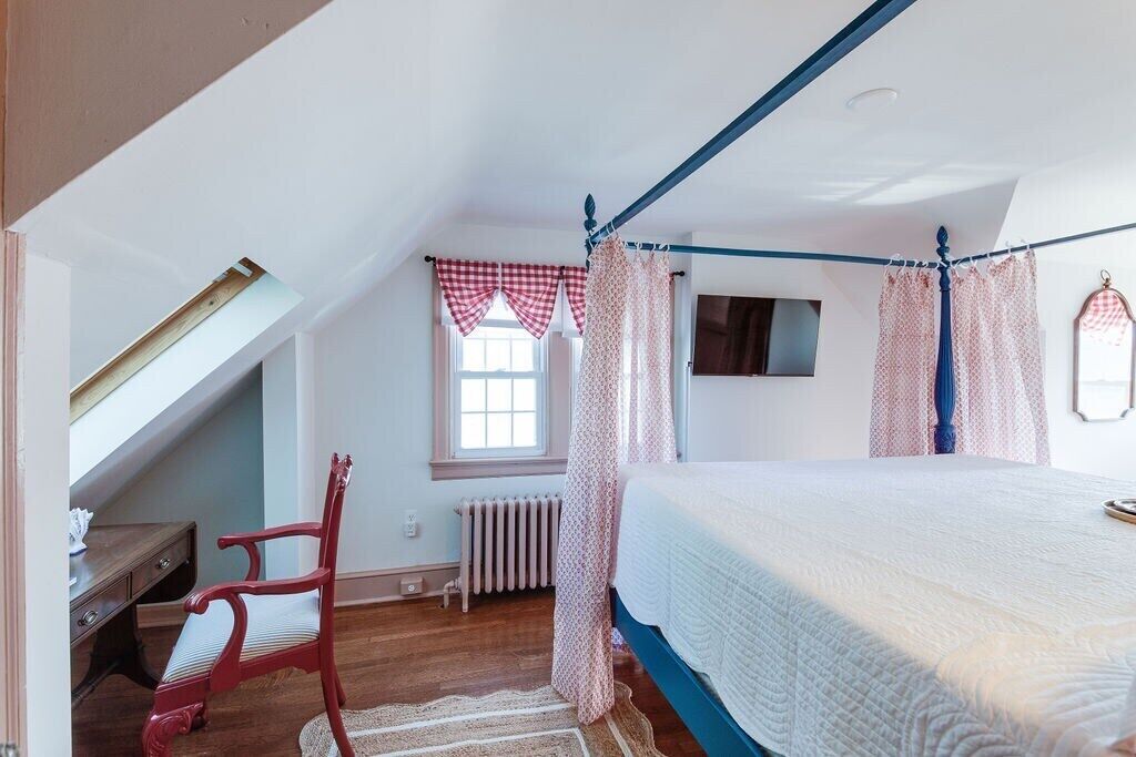 A bedroom with a four-poster bed, a red chair, a desk, and a window with curtains.