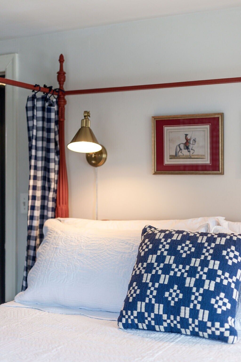 Un lit avec une literie blanche et un oreiller à motifs bleus, une lampe fixée au mur, un tableau encadré et un rideau à carreaux.