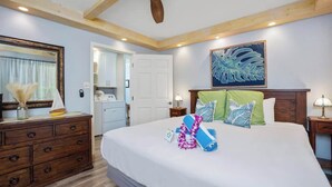 Une chambre à coucher comprenant un lit, une commode en bois, un ventilateur de plafond, des œuvres d’art et une porte donnant accès à une salle de bain.