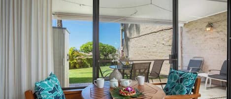 Una mesa redonda de madera con sillas, un paraguas blanco y vistas a un jardín a través de puertas de cristal amplias.