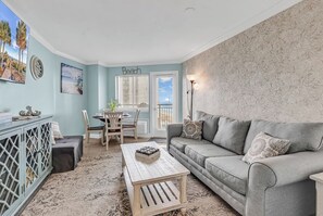 A living room with a grey sofa, a wooden coffee table, a dining table with chairs, and a wall with a 'Beach' sign.