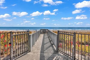A wooden boardwalk leading to a beach with a clear sky and a fence on either side.