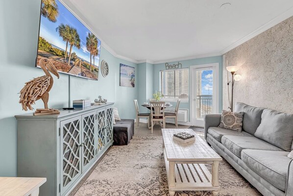A living room with a sofa, a coffee table, a dining area with a table and chairs, a cabinet with a decorative item, and a wall with a beach-themed painting.