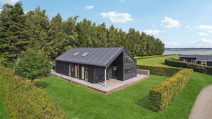 A modern house with a sloped roof and large windows, surrounded by lush greenery and a body of water in the background. - 2 bedroom cozy home in Juelsminde (Juelsminde)