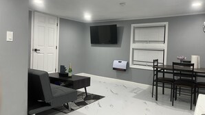 Una sala de estar moderna con un sofá gris, una mesa de comedor con sillas, un televisor de pantalla plana montado en la pared y una ventana con persianas.