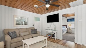 Un salon chaleureux avec un canapé beige, une table basse blanche et un tapis à motifs. Un téléviseur à écran plat est fixé au mur, et une petite table est agrémentée d’objets décoratifs.