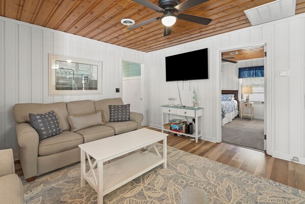 A cozy living room with a beige sofa, a white coffee table, and a patterned rug. A flat-screen TV is mounted on the wall, and there's a small table with decorative items. - Cozy, pet-friendly home - blocks from the ocean - close to shops, eateries, entertainment (North Myrtle Beach)