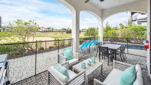 En overdækket terrasse med udendørs møbler, en swimmingpool og udsigt til et boligområde.