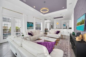 A spacious living room with white leather sofas, a coffee table, and a TV mounted on the wall.