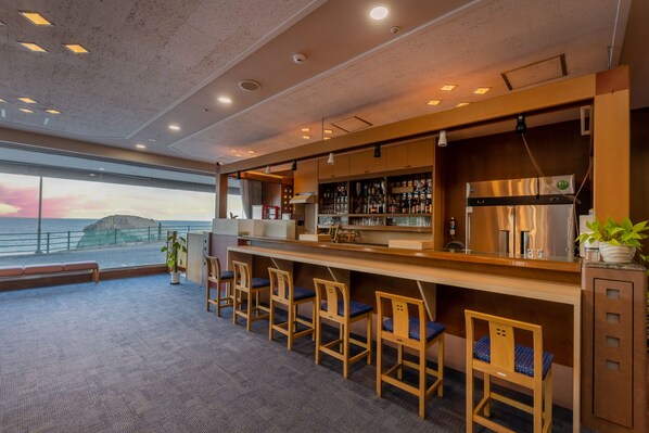 A bar area with a view of the sea, bar stools, and a counter with a refrigerator and shelves.
