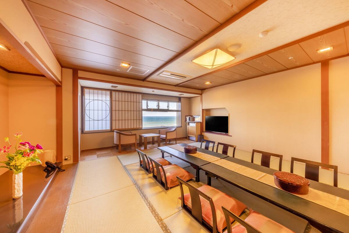 A traditional Japanese-style room with tatami flooring, a long dining table, and a television.