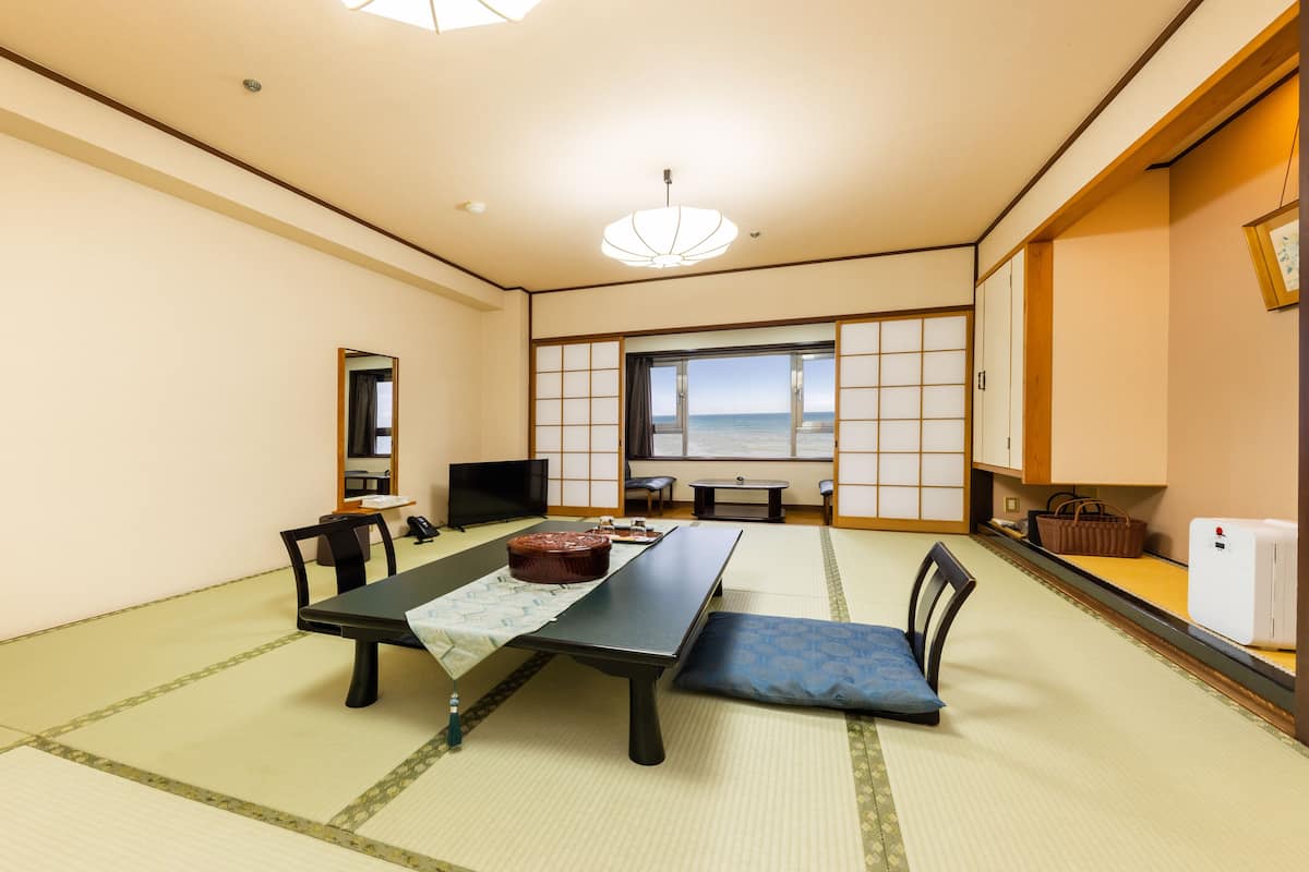 A traditional Japanese room with tatami flooring, a low table with a cushion, a television, and a view of the sea through a shoji screen.
