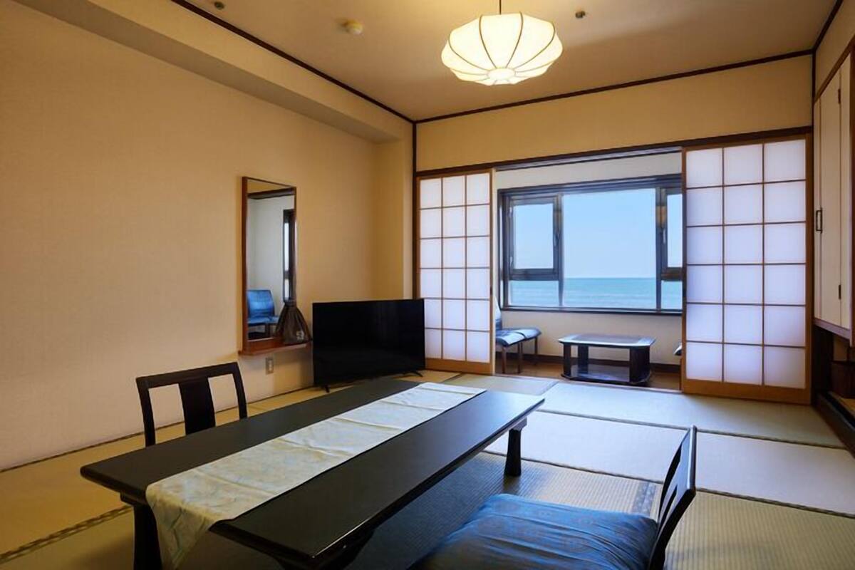 A traditional Japanese room with tatami flooring, a low table, a television, and a view of the sea.