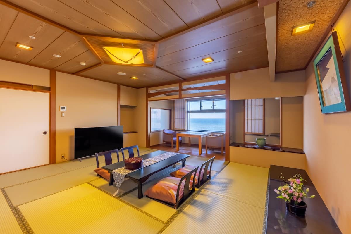 A traditional Japanese-style room with tatami flooring, a low table with cushions, a television, and a view of the sea through large windows.
