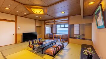 Ein traditioneller japanischer Raum mit Tatami-Boden, einem niedrigen Tisch mit Kissen, einem Fernseher und Meerblick durch große Fenster.