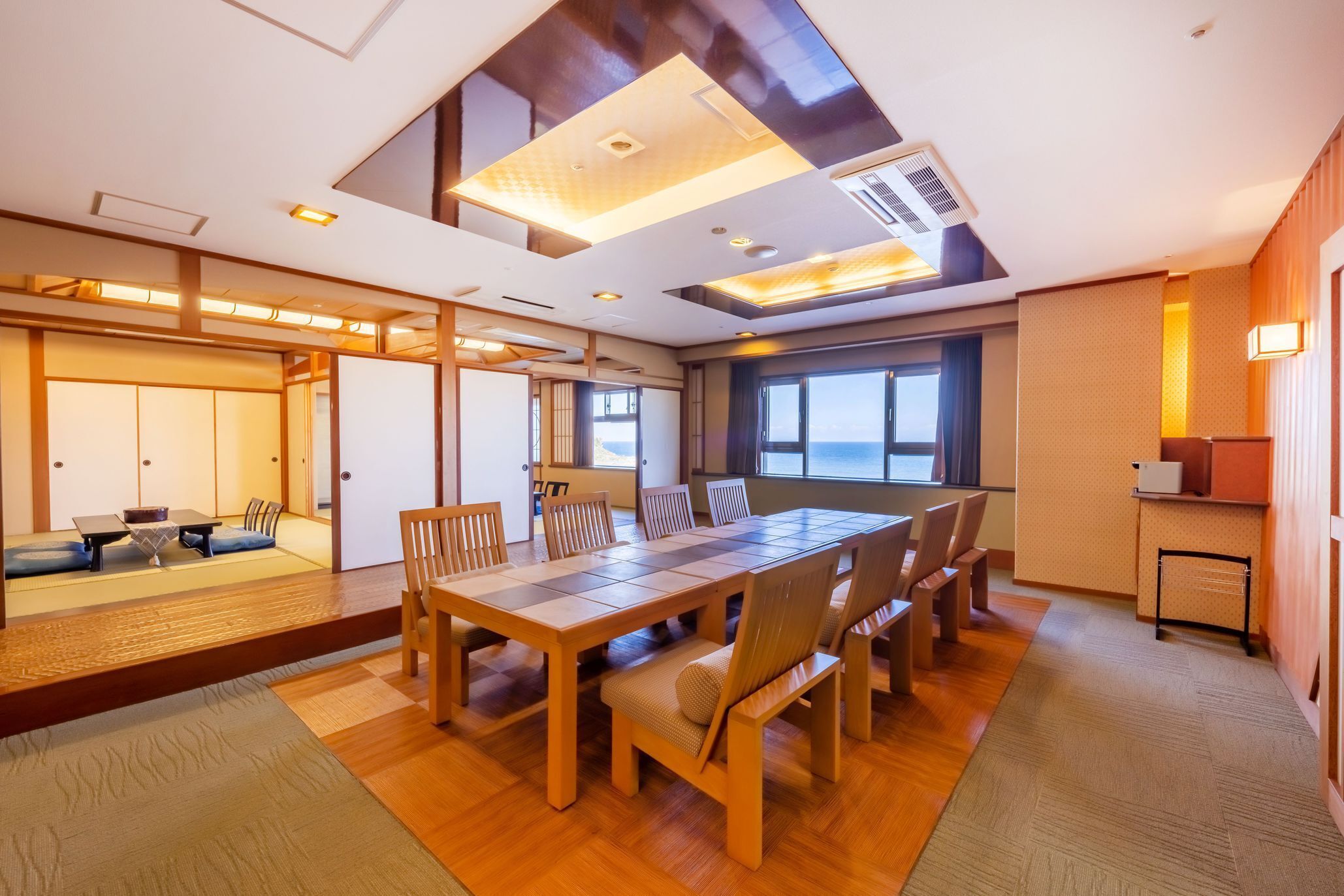 A modern hotel room with a wooden dining table, chairs, a sofa, and a view of the sea.